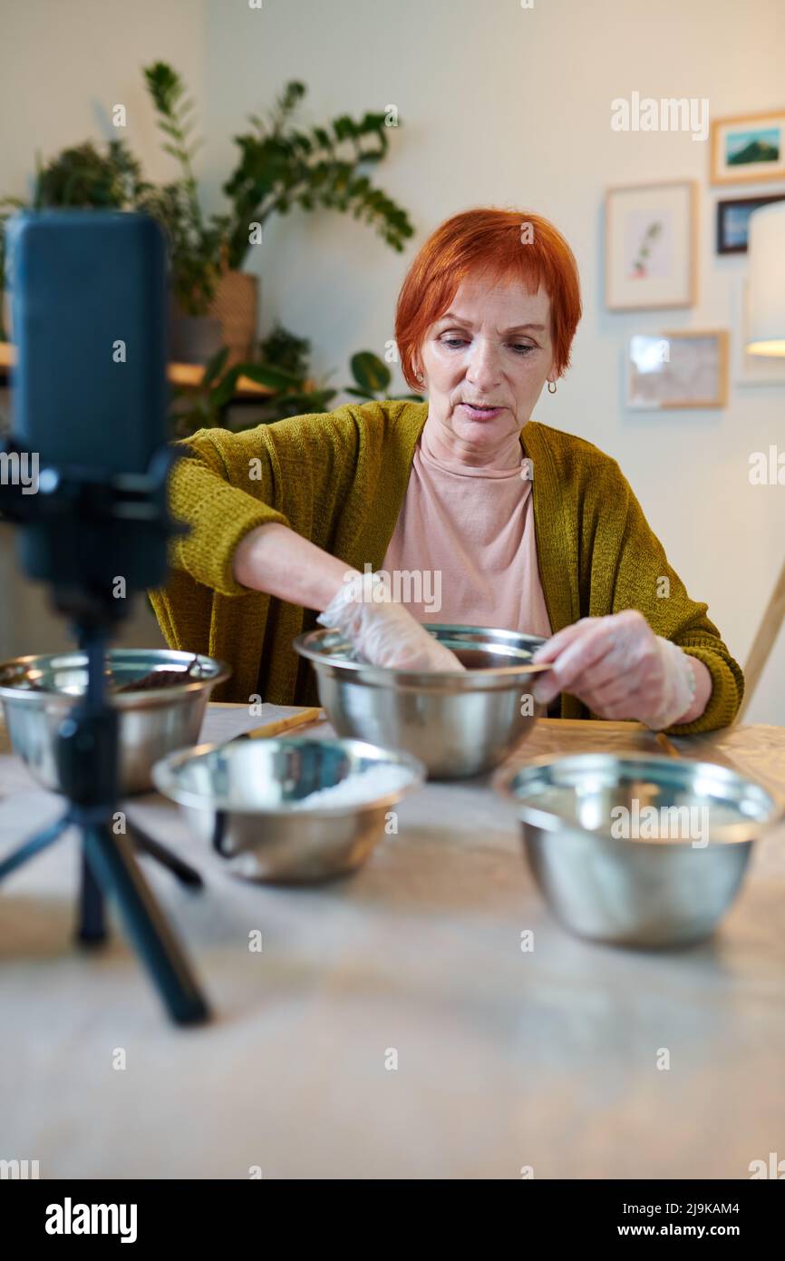 Mature woman sitting at table with bowls and talking about planting and gardening during live broadcasting on mobile phone Stock Photo