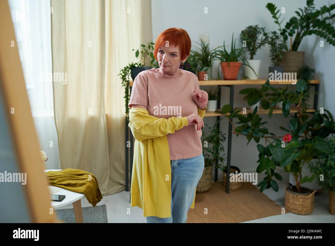 Elderly elegant woman standing in front of mirror and admiring herself while trying on new cardigan in room with house plants in background Stock Photo