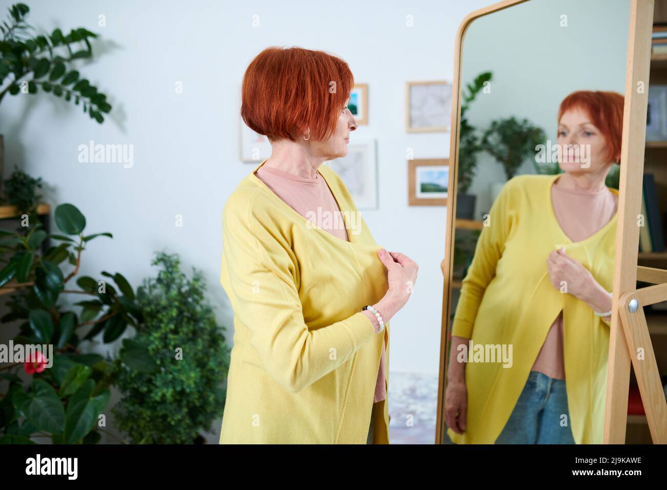 Senior woman with red hair looking at her reflection in mirror while trying on new yellow cardigan Stock Photo