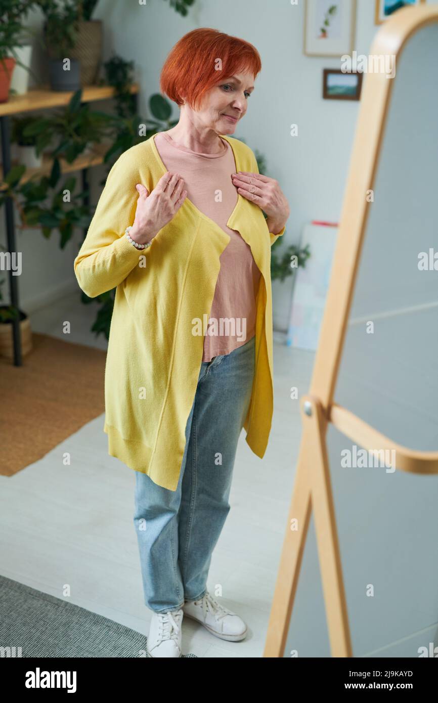 Senior woman wearing new yellow cardigan looking at herself in mirror standing in room Stock Photo