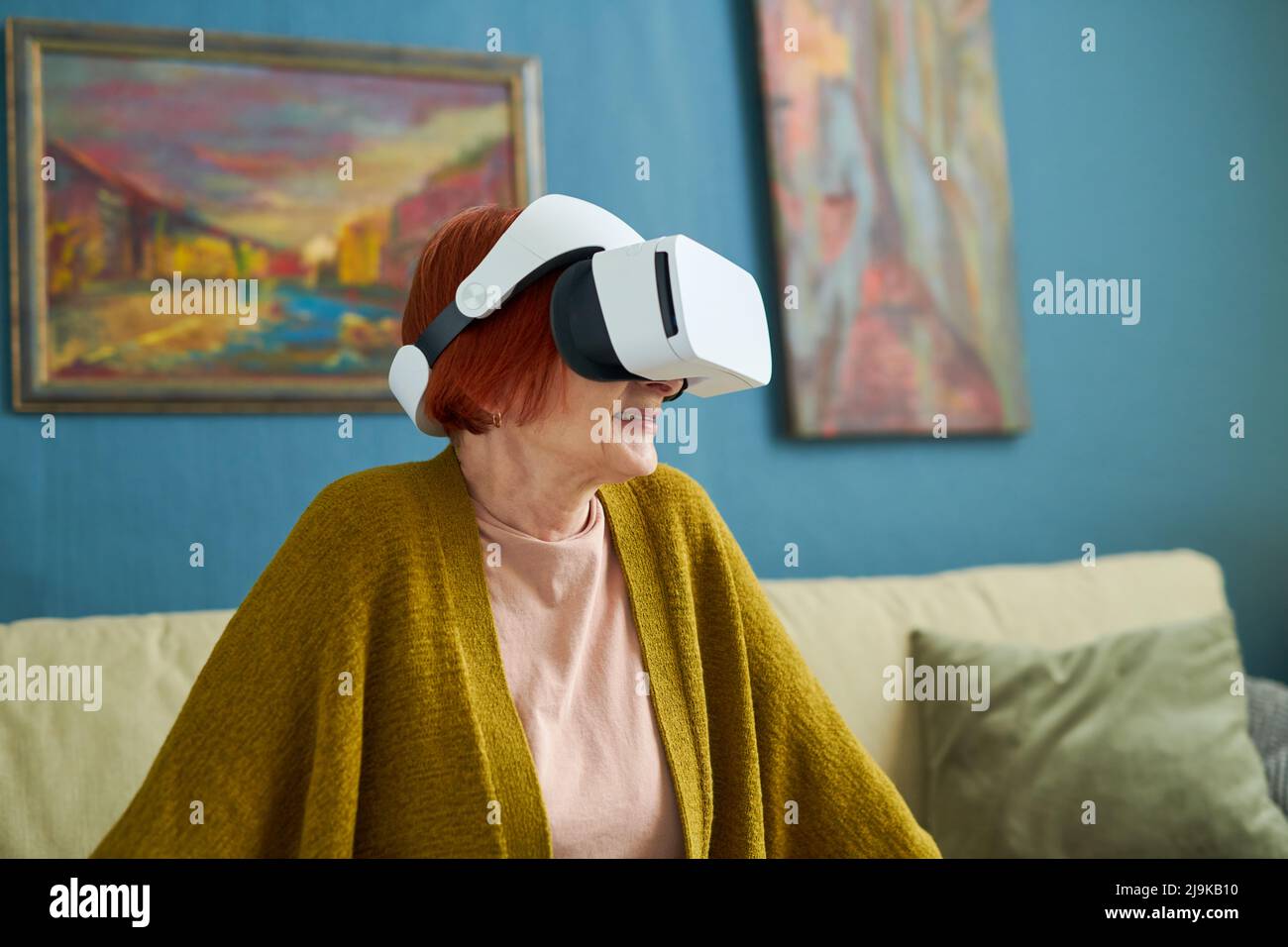 Grandmother in virtual reality goggles playing online while resting on sofa in room Stock Photo