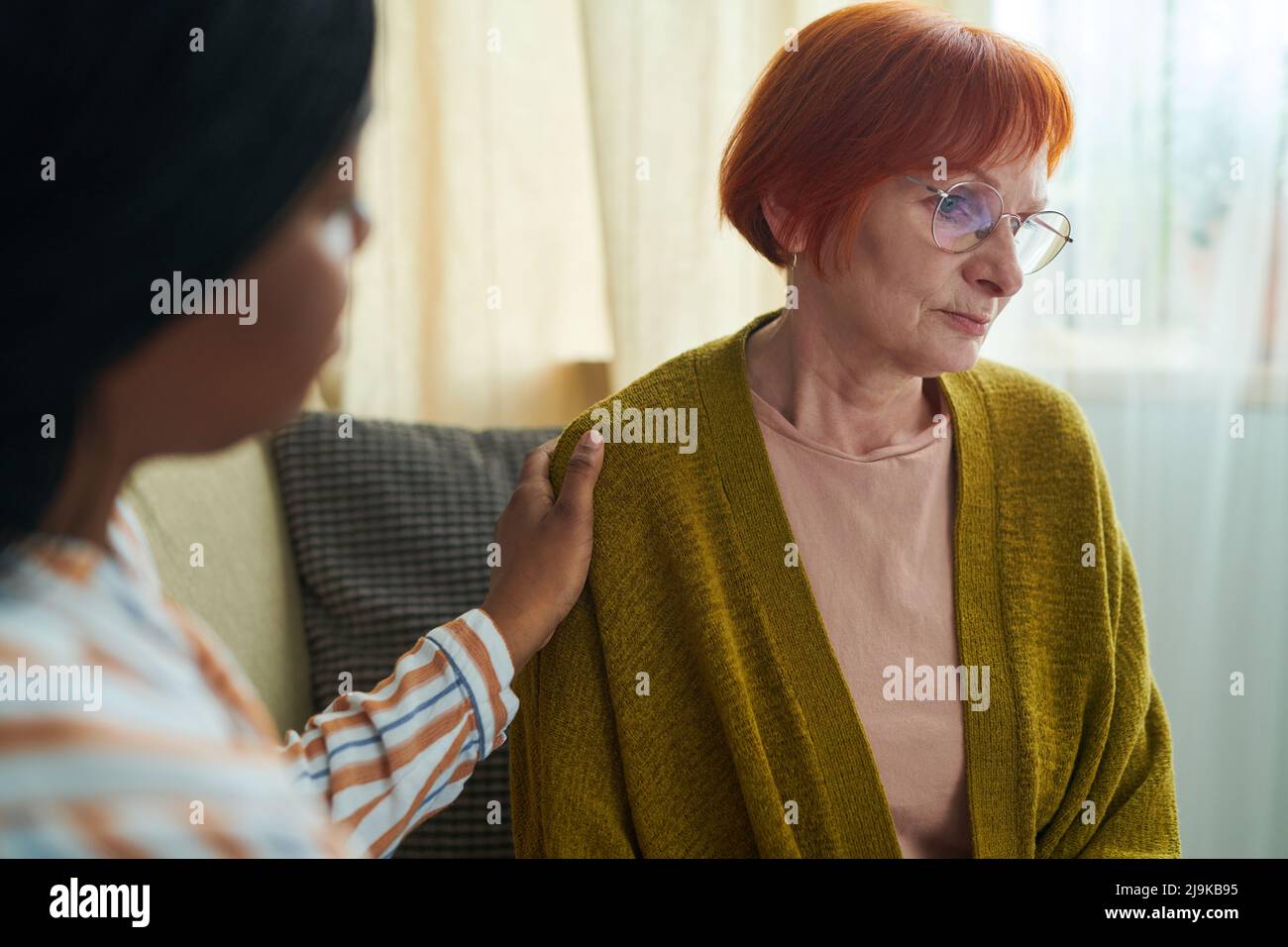 African caregiver supporting senior woman in eyeglasses with sad expression while sitting on sofa Stock Photo