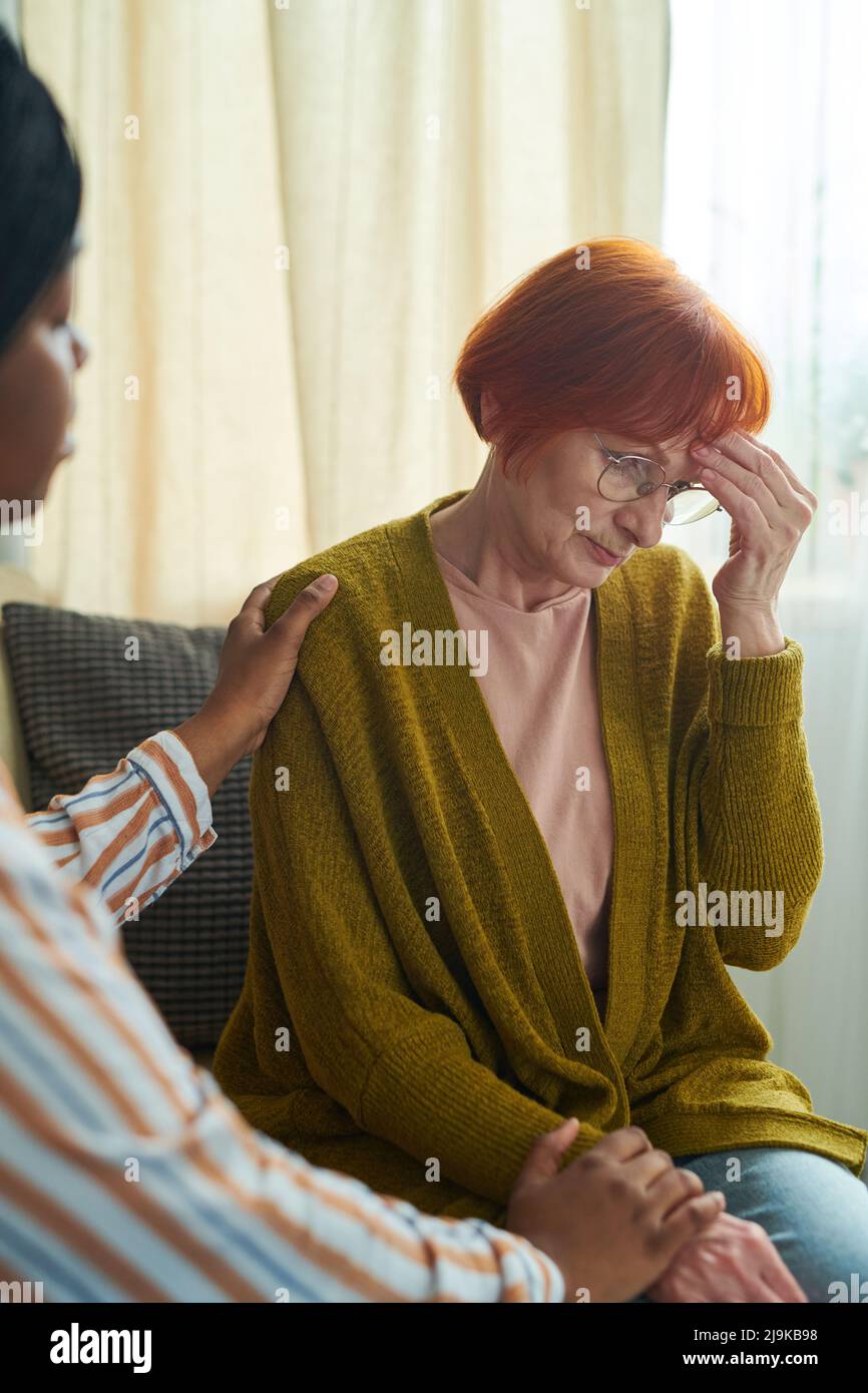 Elderly woman in eyeglasses sitting on sofa in depression while volunteer supporting her Stock Photo