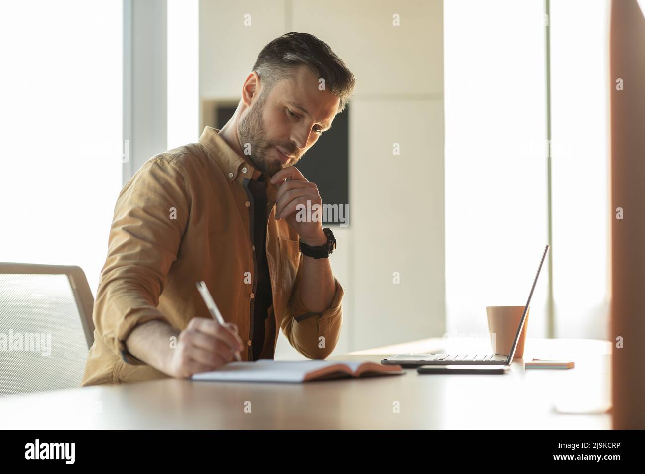 Thoughtful Businessman Writing Taking Notes Using Laptop At Workplace Stock Photo