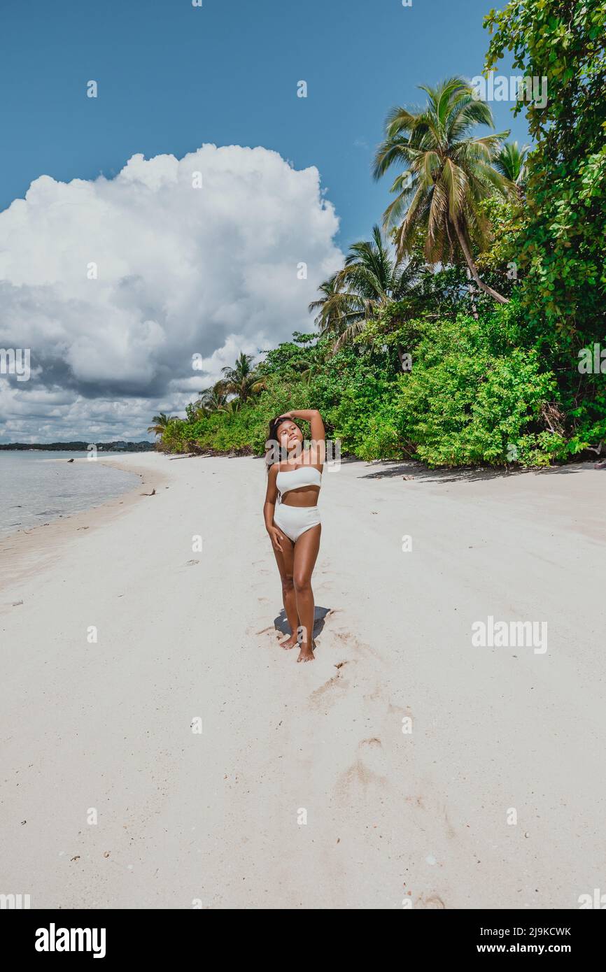 Young tan asian female in a white bikini on a white sand tropical island beach surrounded by palm coconut trees on a tropical sunny day Stock Photo