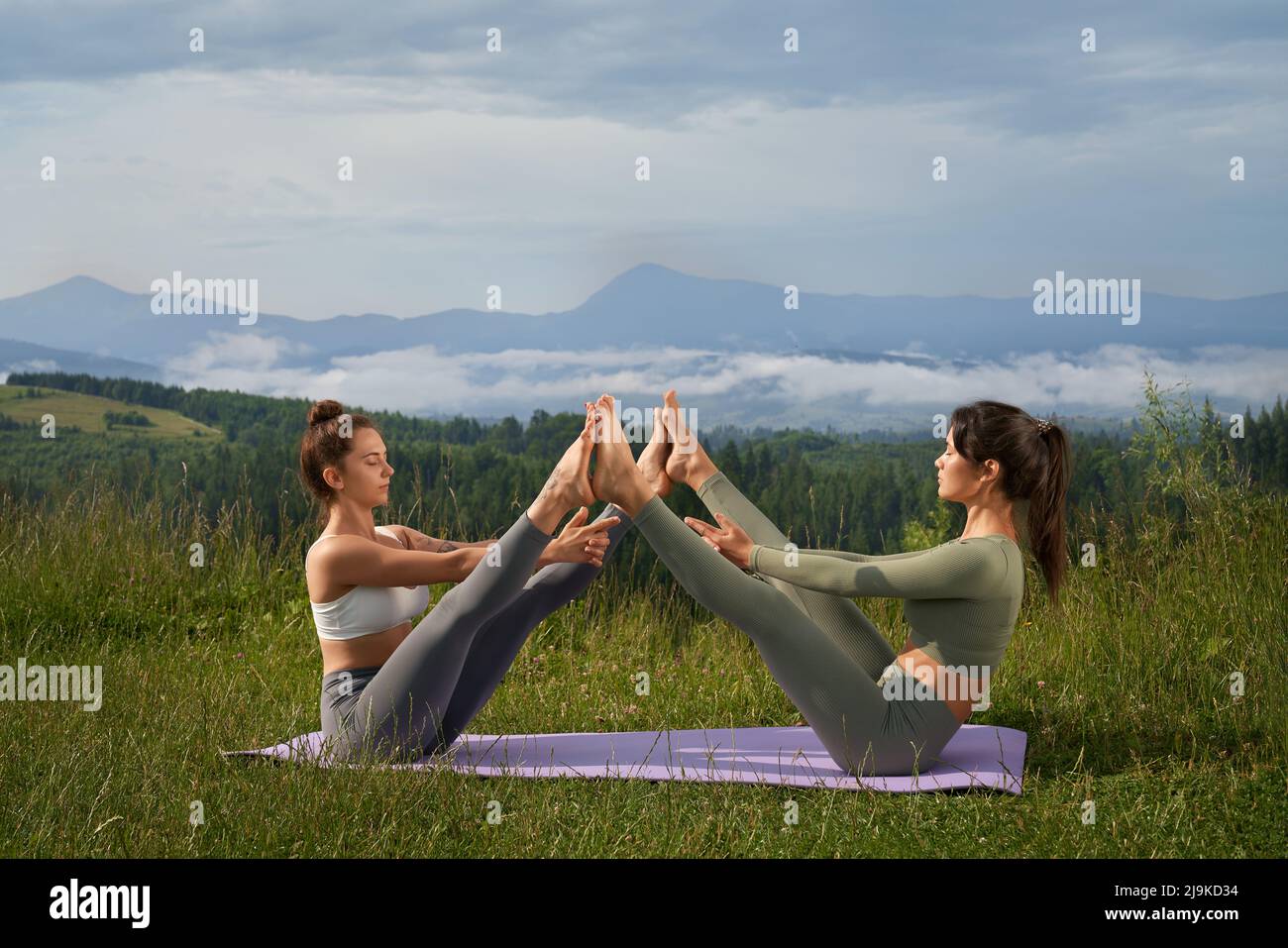 Young caucasian women stretching legs and meditating outdoors. Two friends sitting on yoga mat with amazing view on summer mountains on background. Stock Photo