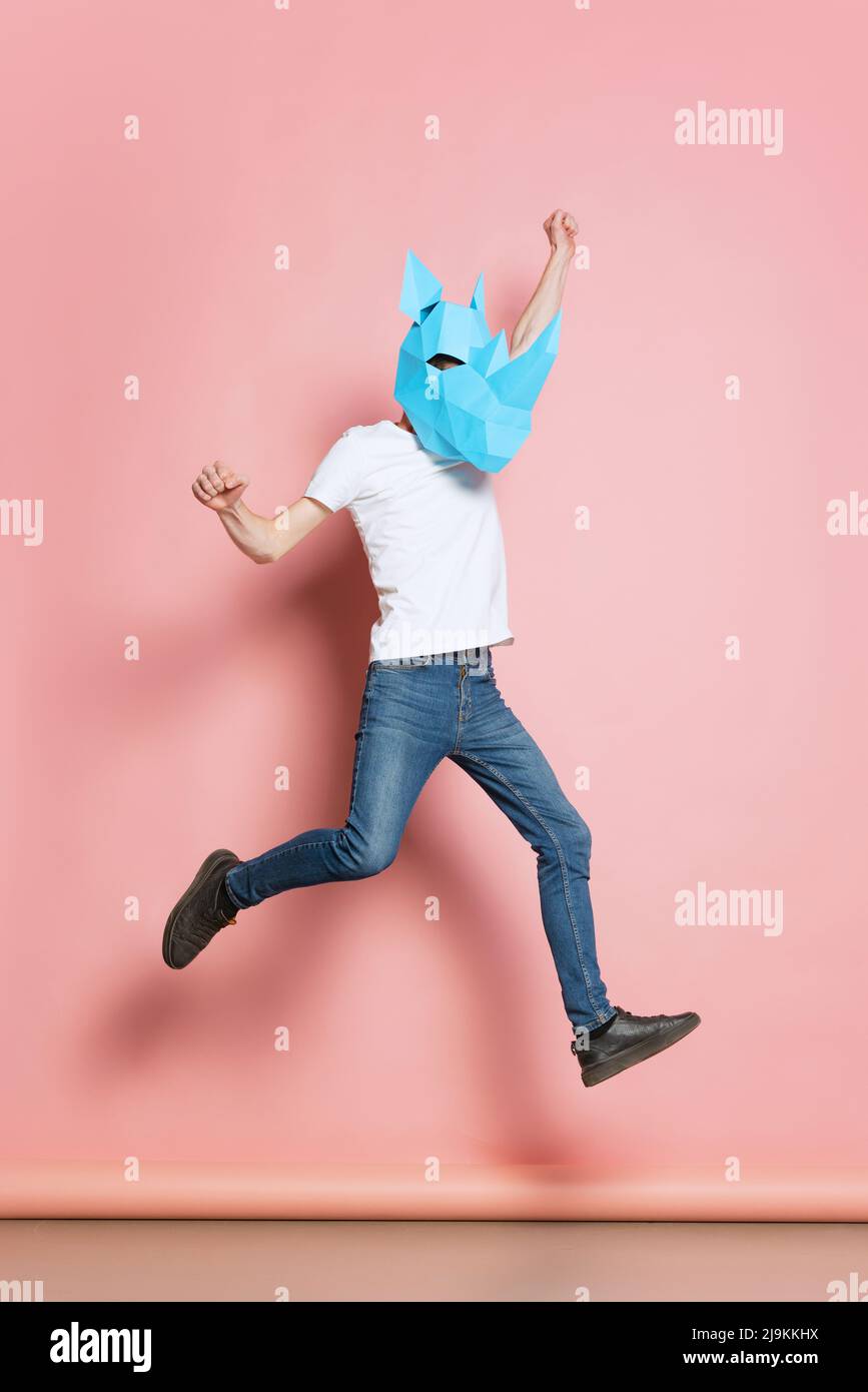 Creative portrait of young man in white t-shirt with cardboard animal mask on his head jumping isolated on pink background. Stock Photo