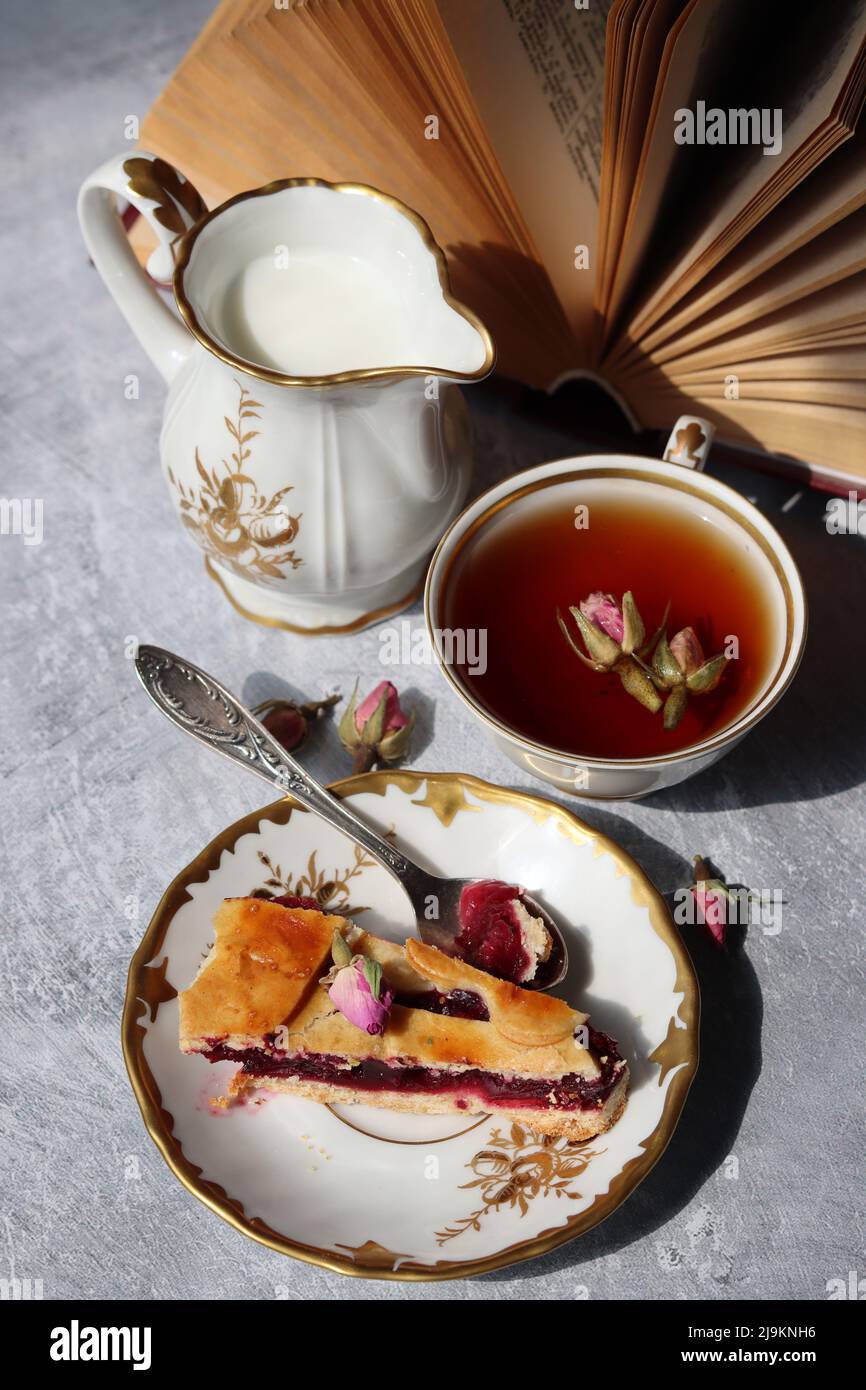 Still life with cup of tea,  jug of milk, cherry pie, small rose flowers. White vintage tableware on a table.  Sweet breakfast close up photo. Stock Photo