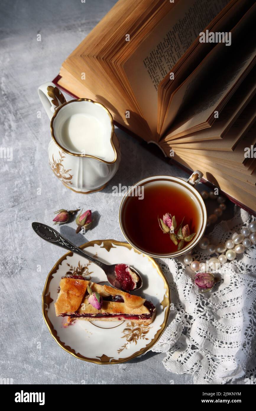 Still life with cup of tea,  jug of milk, cherry pie, small rose flowers. White vintage tableware on a table.  Sweet breakfast close up photo. Stock Photo