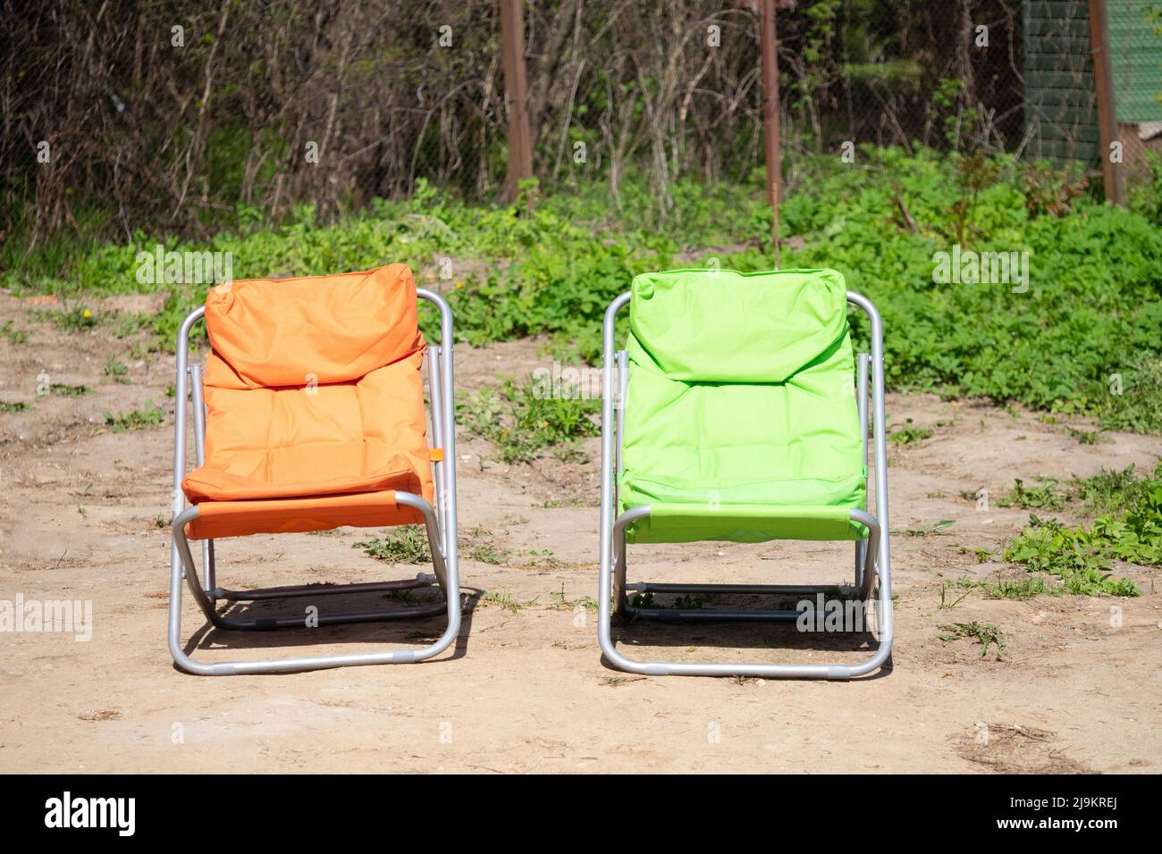 Orange and green folding tourist chair for leisure on the background of brown earth, green grass, iron mesh fence under sun Stock Photo