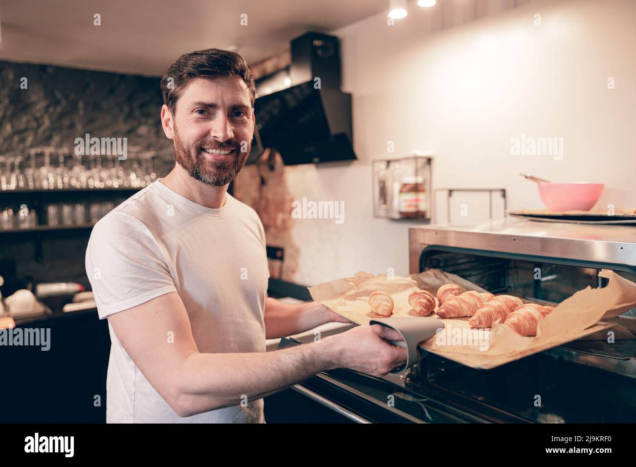 Caucasian attractive male baker taking out croissants out of oven and smiling. Bakery kitchen. Stock Photo