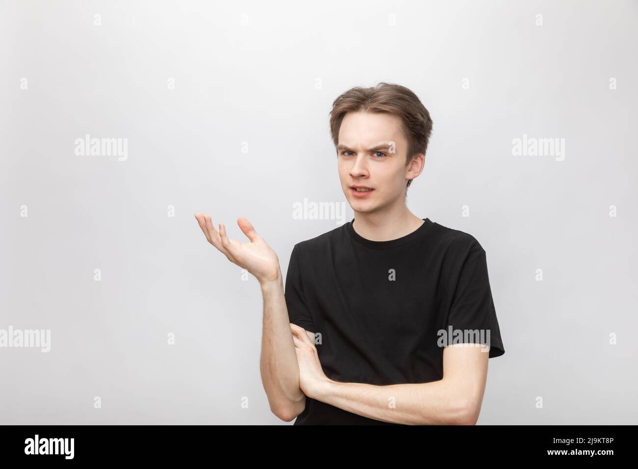 Portrait of doubtful young man wearing black tshirt holding hand in questioning gesture looking at camera suspiciously. Studio shot on gray background Stock Photo