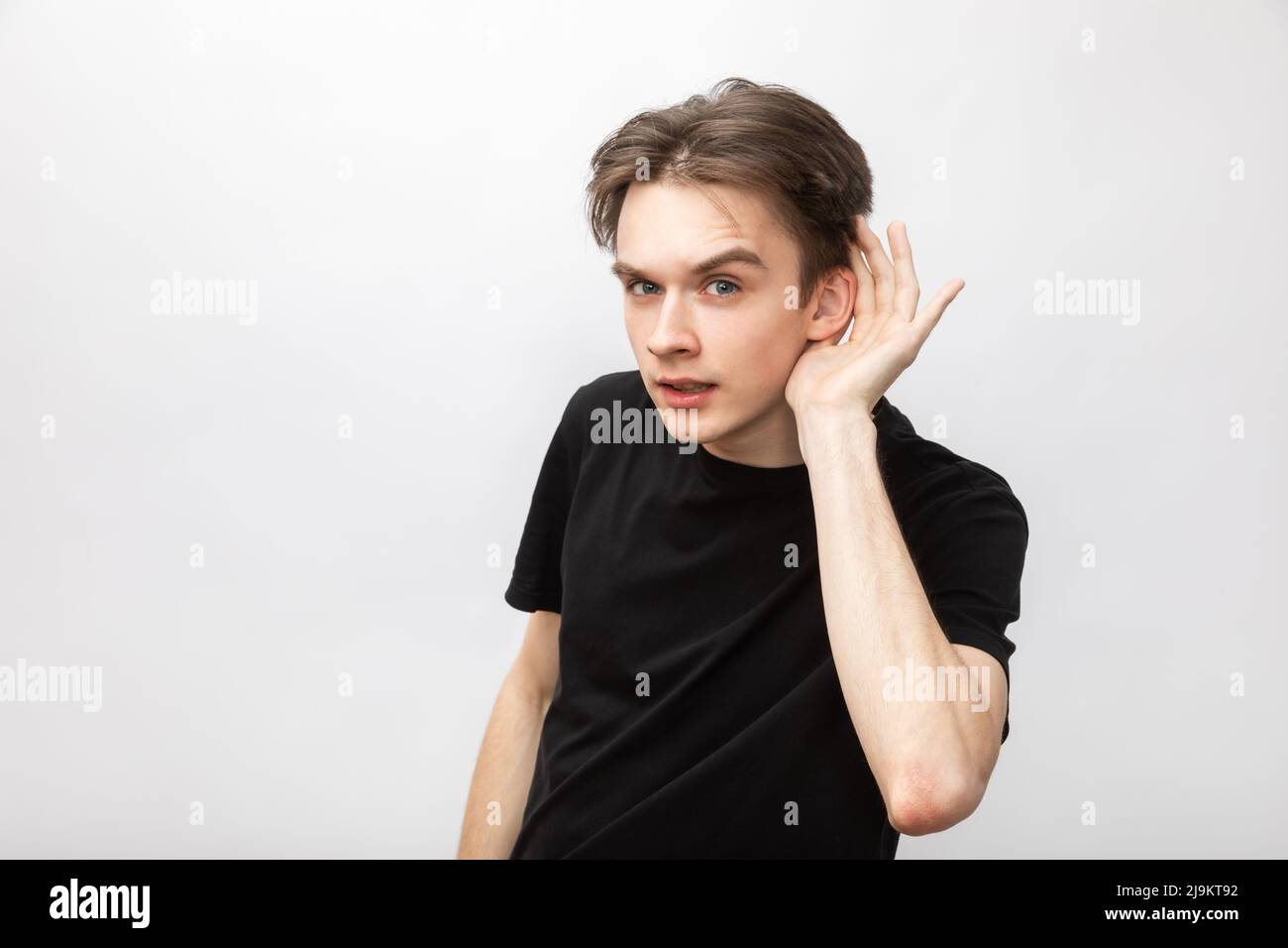 Portrait of young man wearing black tshirt listening holding hand near ear looking at camera. Studio shot on gray background Stock Photo