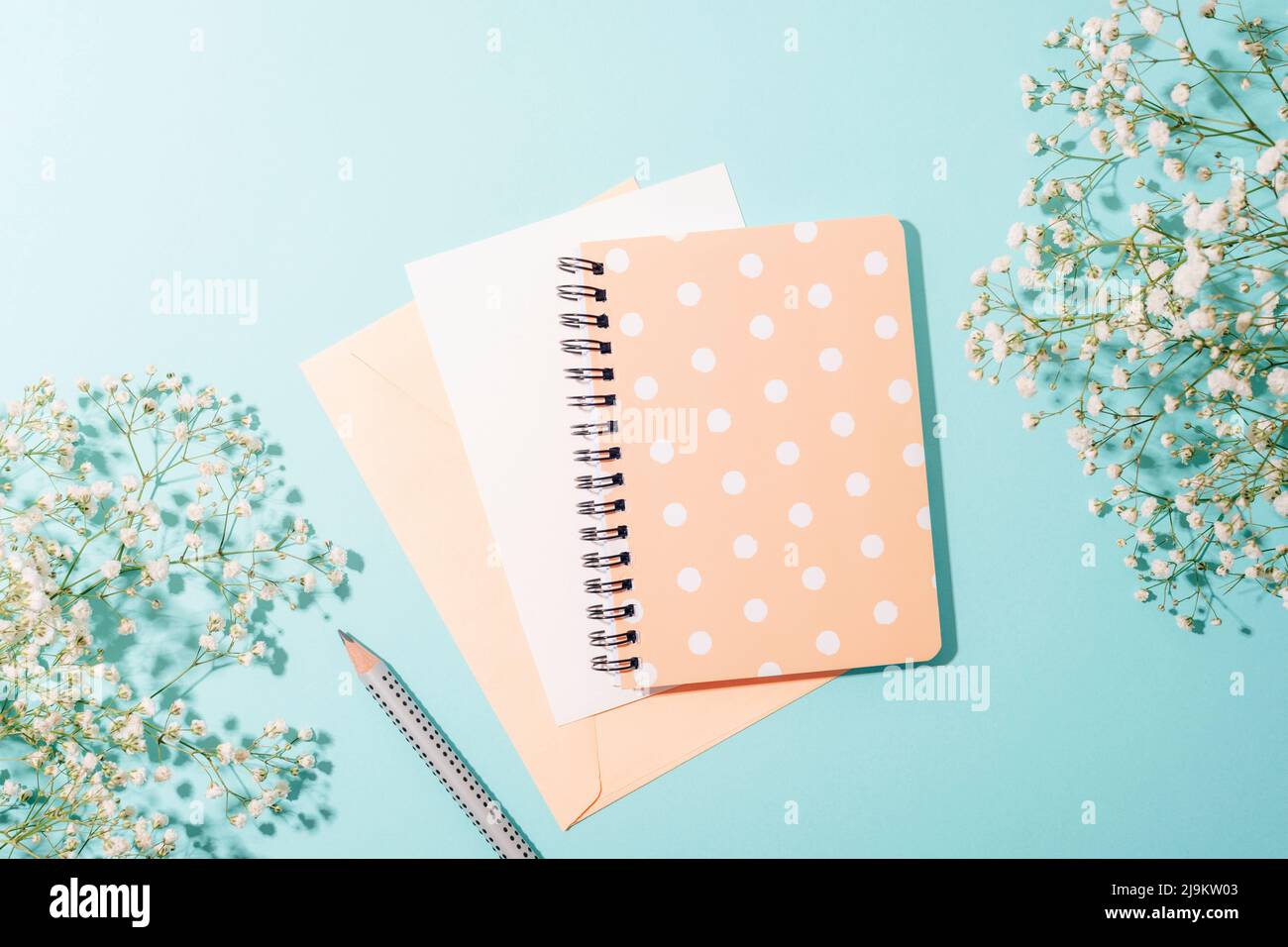 Notepad and envelope with blank sheet, pencil on blue background with gypsophila flowers in sunlight. Top view, flat lay. Stock Photo