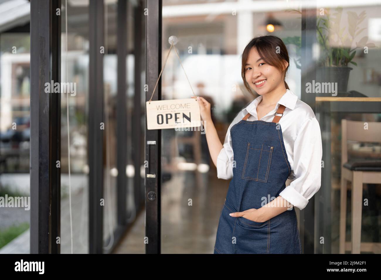 Young Asia manager girl changing a sign from closed to open sign on door cafe looking outside waiting for clients after lockdown. Owner small business Stock Photo