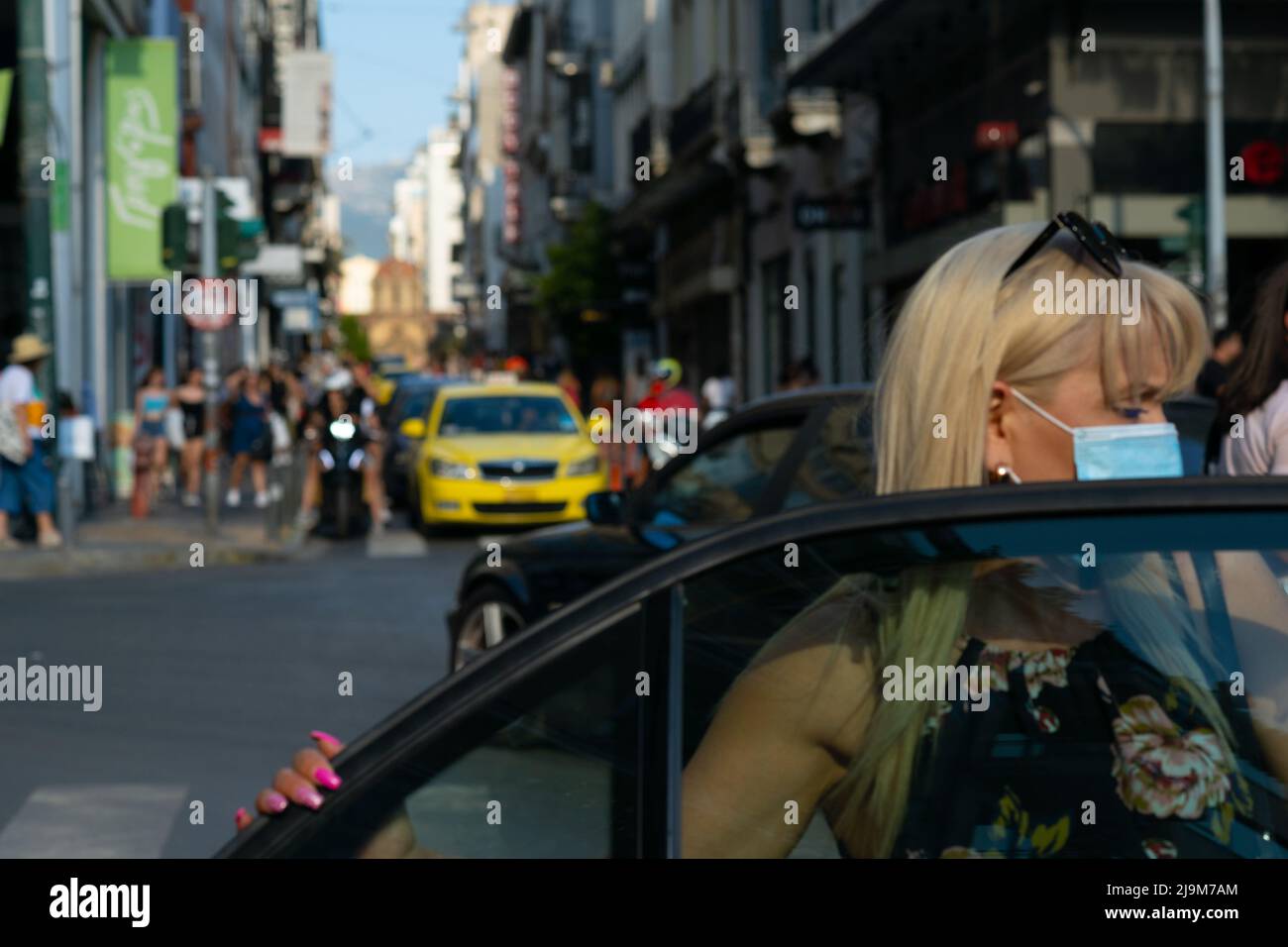 Lady wearing a Covid mask steps into a car on a busy street in Athens Stock Photo