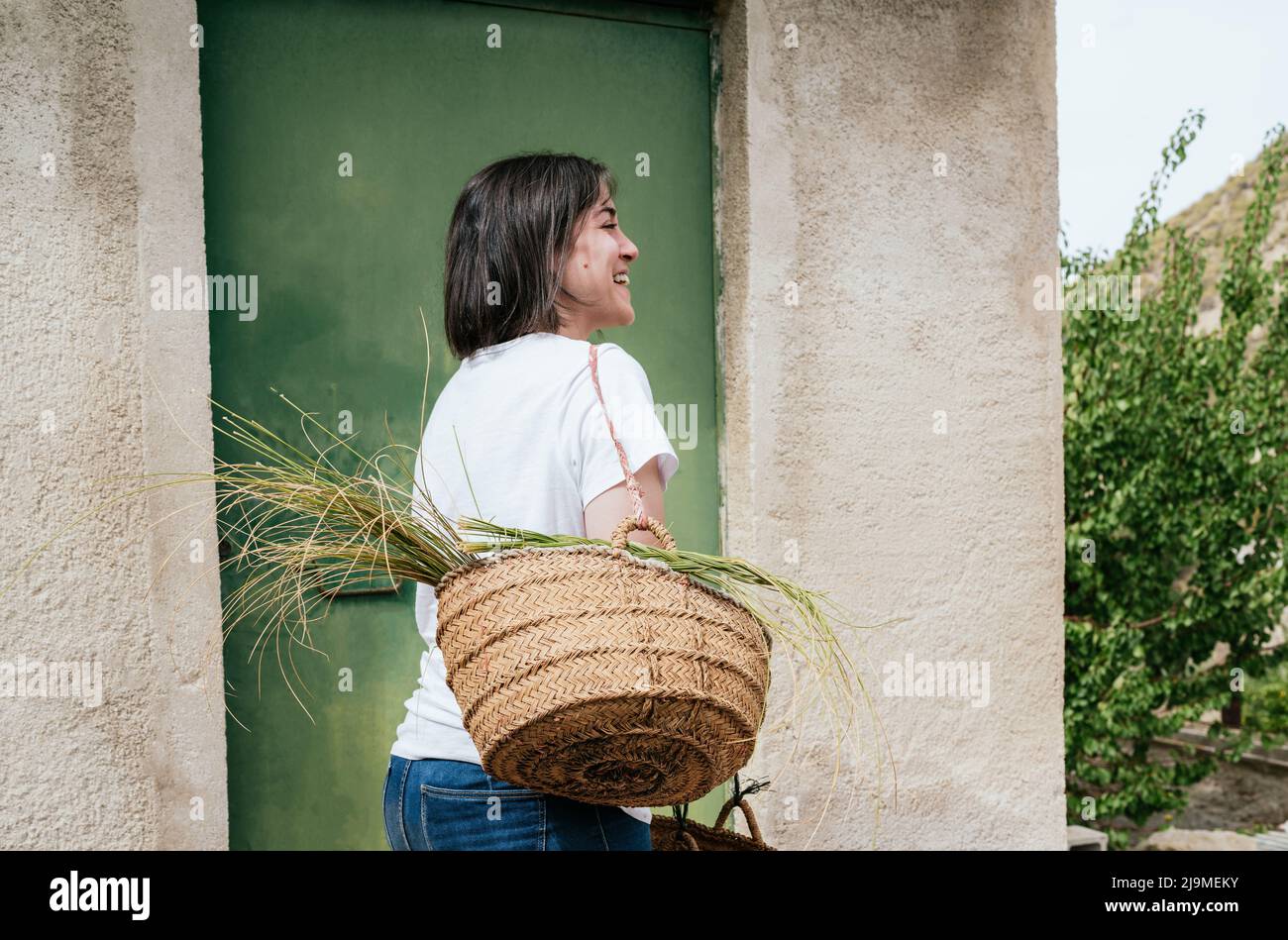 Side view of cheerful young Hispanic female farmer with dark hair in casual clothes holding handmade wicker basket with esparto grass and smiling in c Stock Photo
