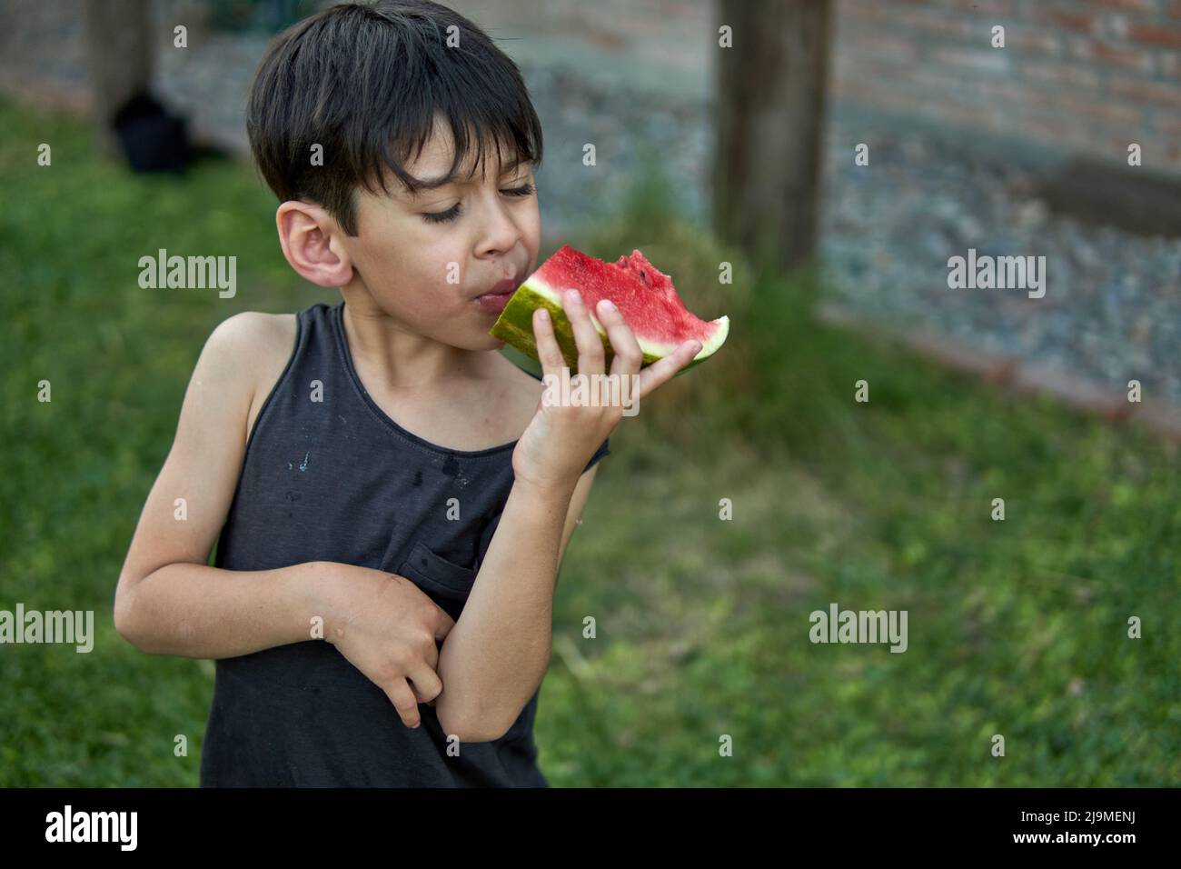 brown latino boy eating watermelon in the garden on a summer afternoon. healthy childhood habit. Selective focus and copy space. Stock Photo