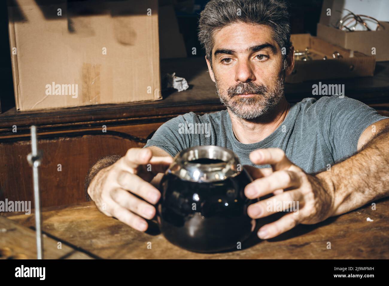 Middle aged bearded male artisan putting handmade silver rim on black ceramic cup placed on table while working in workshop Stock Photo