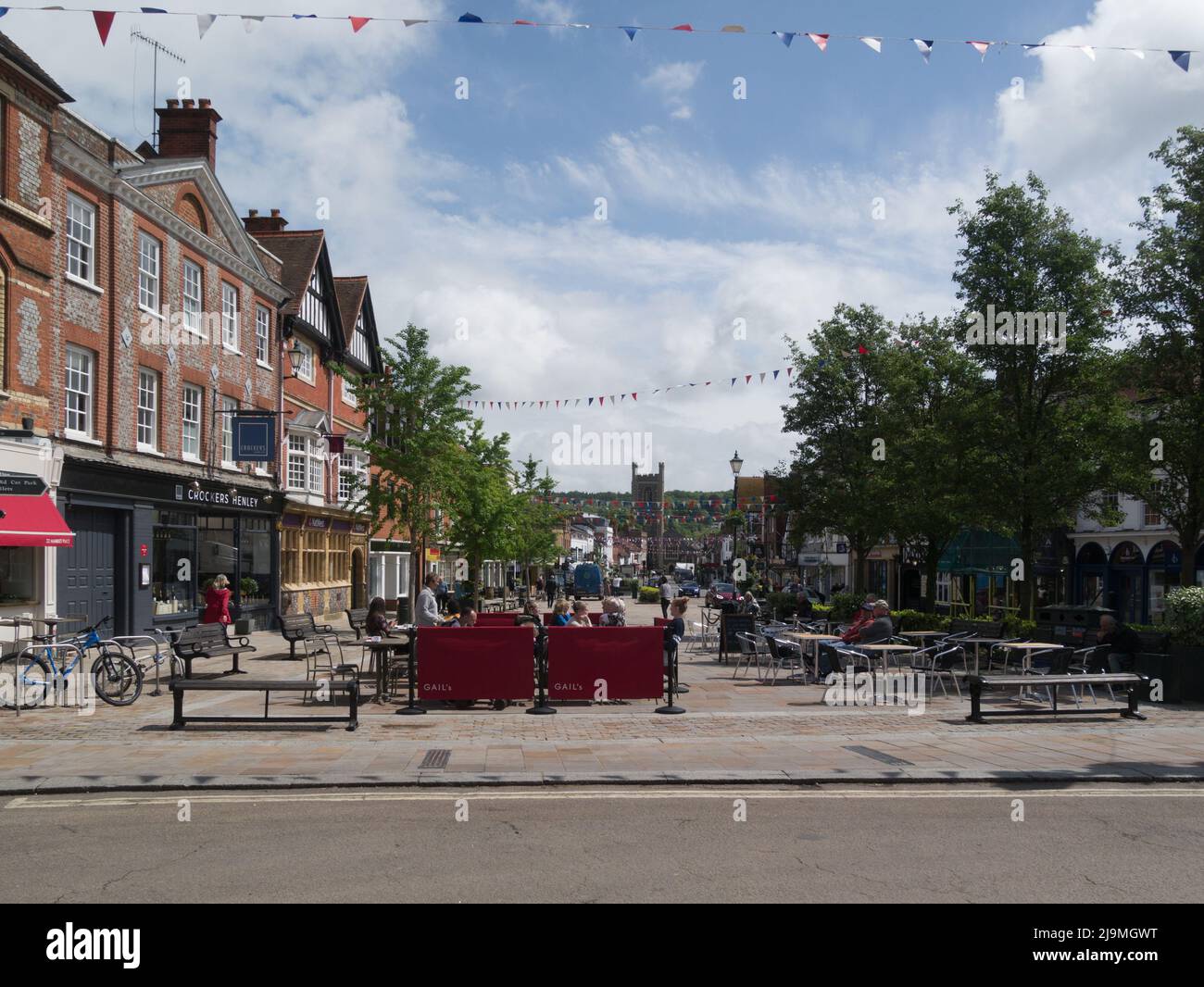 People sitting out in sunshine in pedestrianised square Henley-on-Thames Oxfordshire England UK view down main street to St Mary's Church on lovely Ma Stock Photo