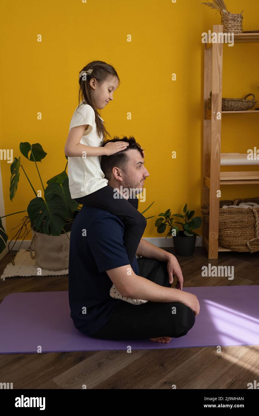 Side view of happy girl smiling and rumpling hair of glad father while sitting on shoulders of adult man after yoga session at home Stock Photo