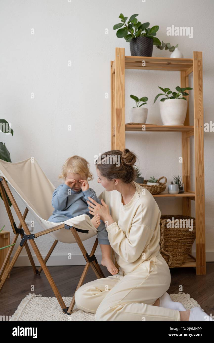 Young female in casual clothes sitting on floor and calming adorable little kid crying and rubbing eyes at home Stock Photo