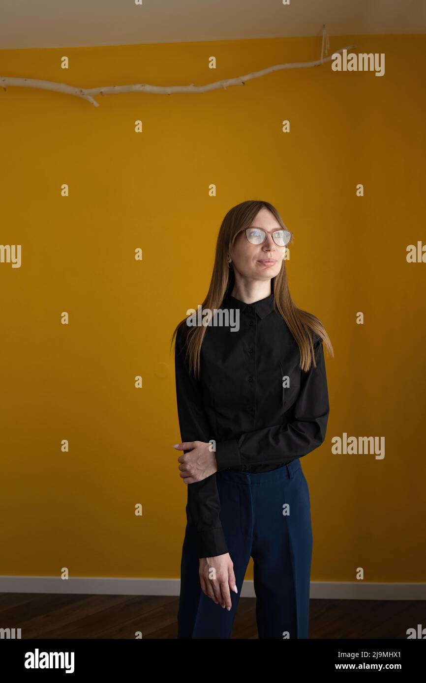 Adult female in smart casual clothes and glasses touching arm and looking away while standing against yellow wall at home Stock Photo