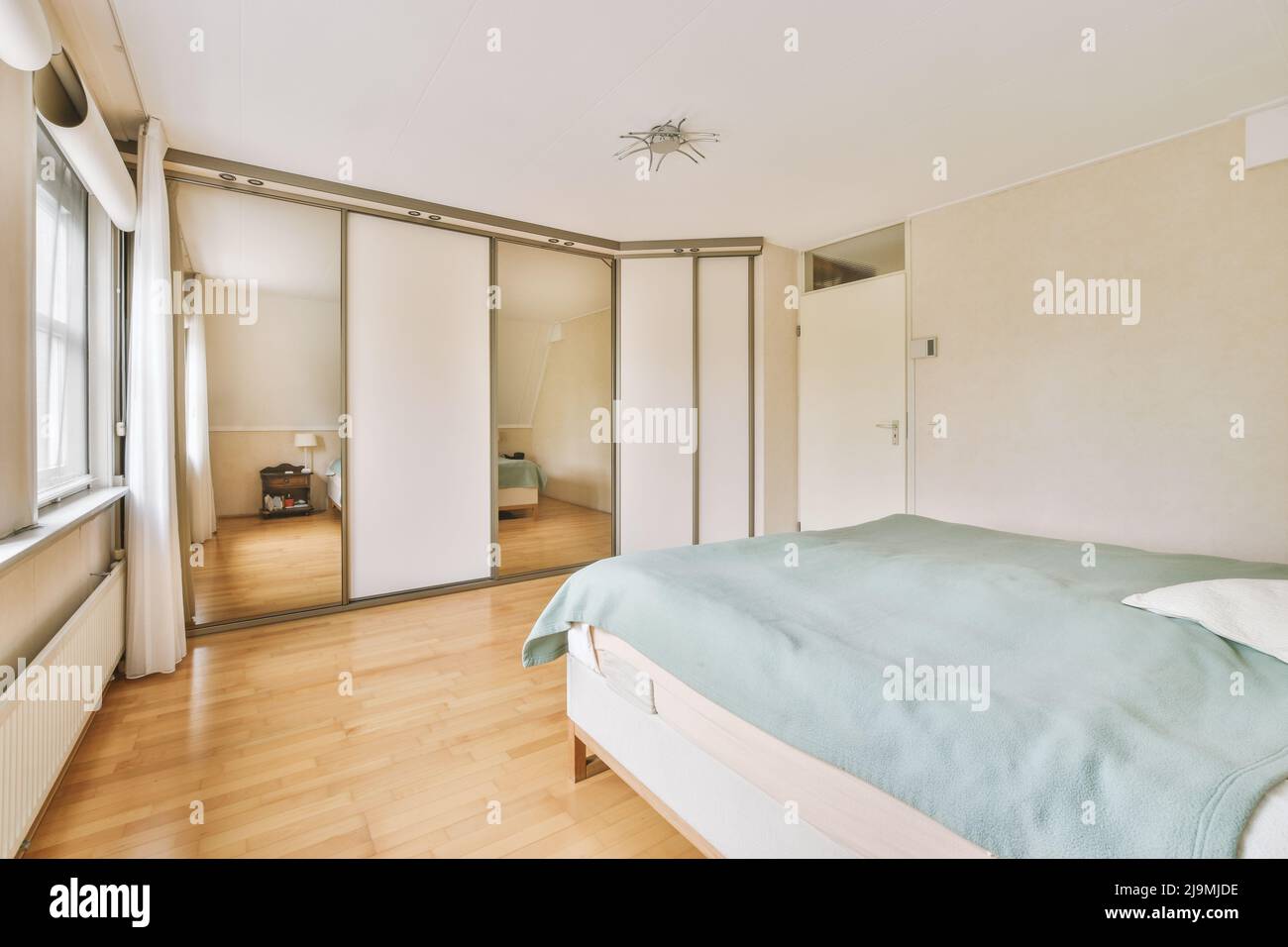 Comfortable bed with blanket placed near glass mirror wardrobe in light bedroom at home Stock