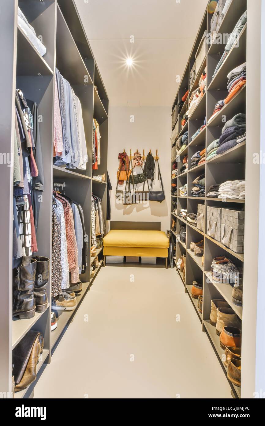 Stylish bags hanging on wall over comfortable pouf in spacious dressing room with closet with various clothes on racks and shelves Stock Photo
