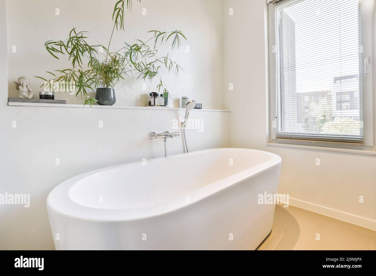 Oval white ceramic bathtub placed near window with jalousie in spacious