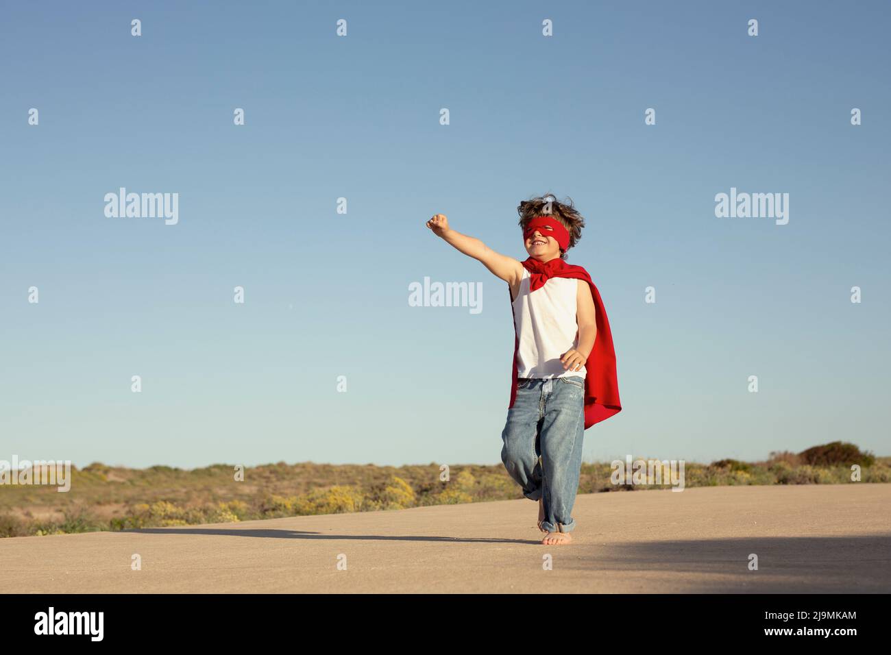 Adorable delighted little child with clenched fists in red superhero cape and mask smiling and looking away while standing against cloudless blue sky Stock Photo