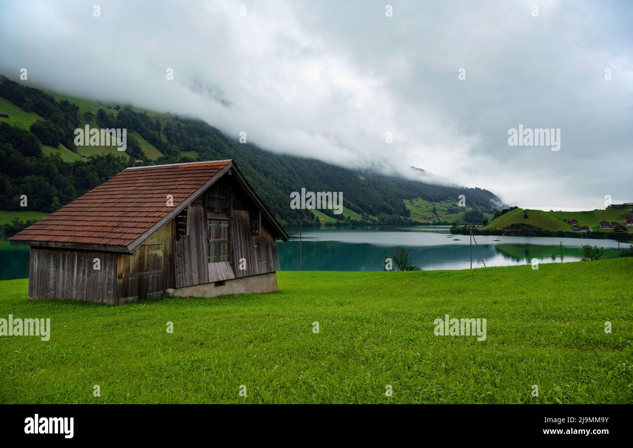 View of a red wooden chalet farm house surrounded by mountains and green meadows, alongside the lake Lungern situated at the alpine village of Lungern Stock Photo