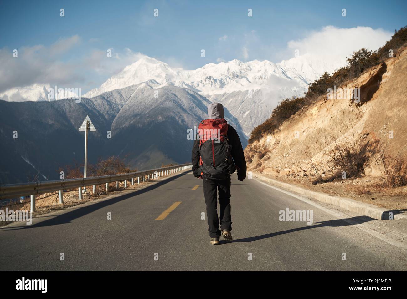 rear view of an asian hiker with backpack walking on highway with snowcapped meili mountain in background Stock Photo