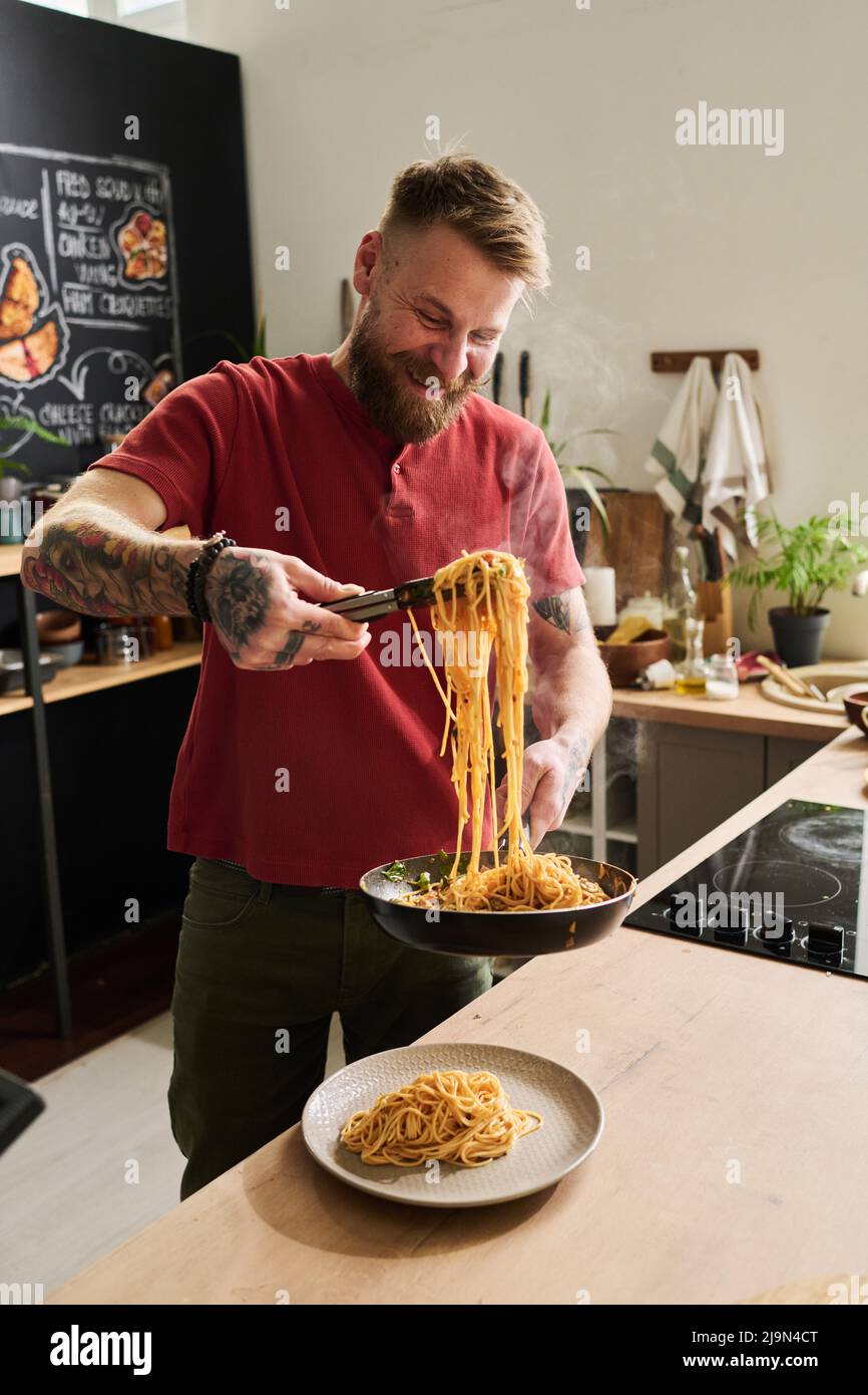Joyful Caucasian chef standing in loft kitchen serving hot spaghetti with kitchen tongues on plate Stock Photo