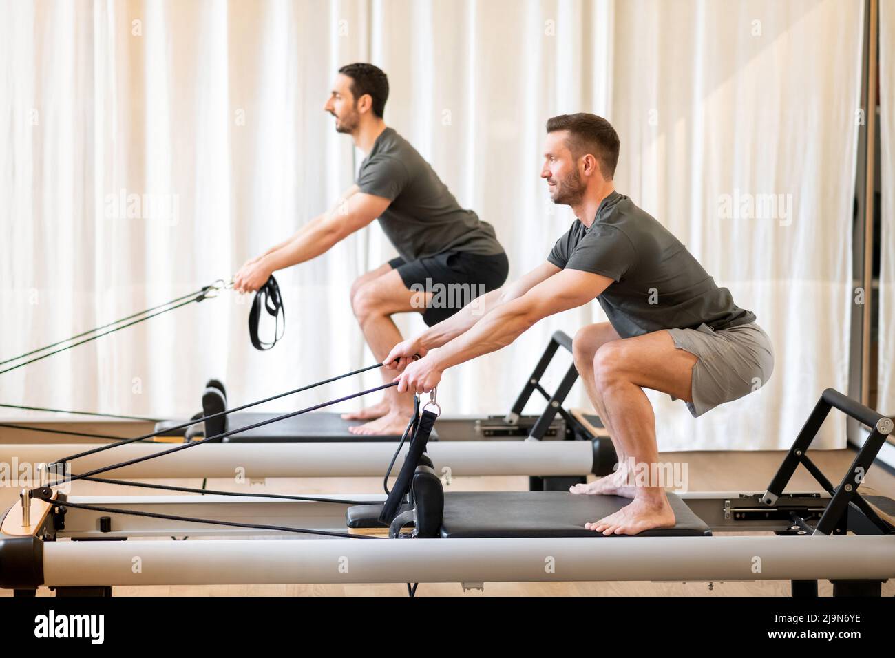 Side view of concentrated sportsmen doing squat exercise while standing on pilates reformers in spacious fitness studio Stock Photo