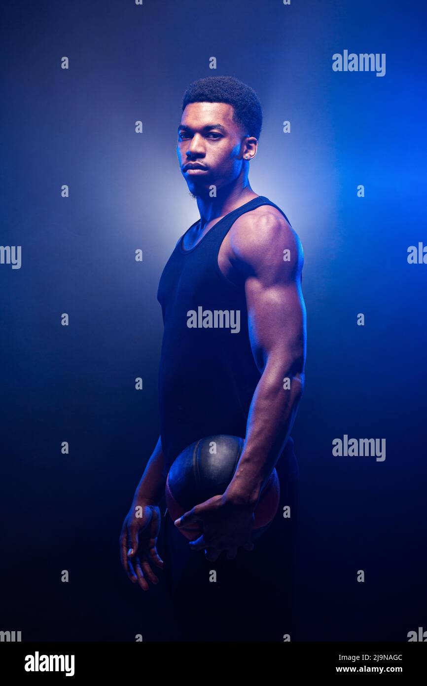 Basketball player lit with blue color holding a ball against smoke background. Serious concentrated african american man. Stock Photo