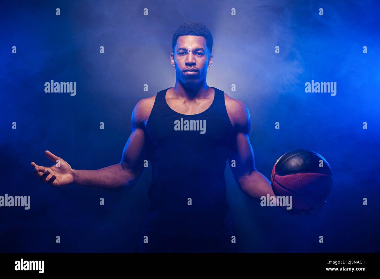 Basketball player lit with blue color posing with a ball against smoke background. Serious concentrated african american man. Stock Photo
