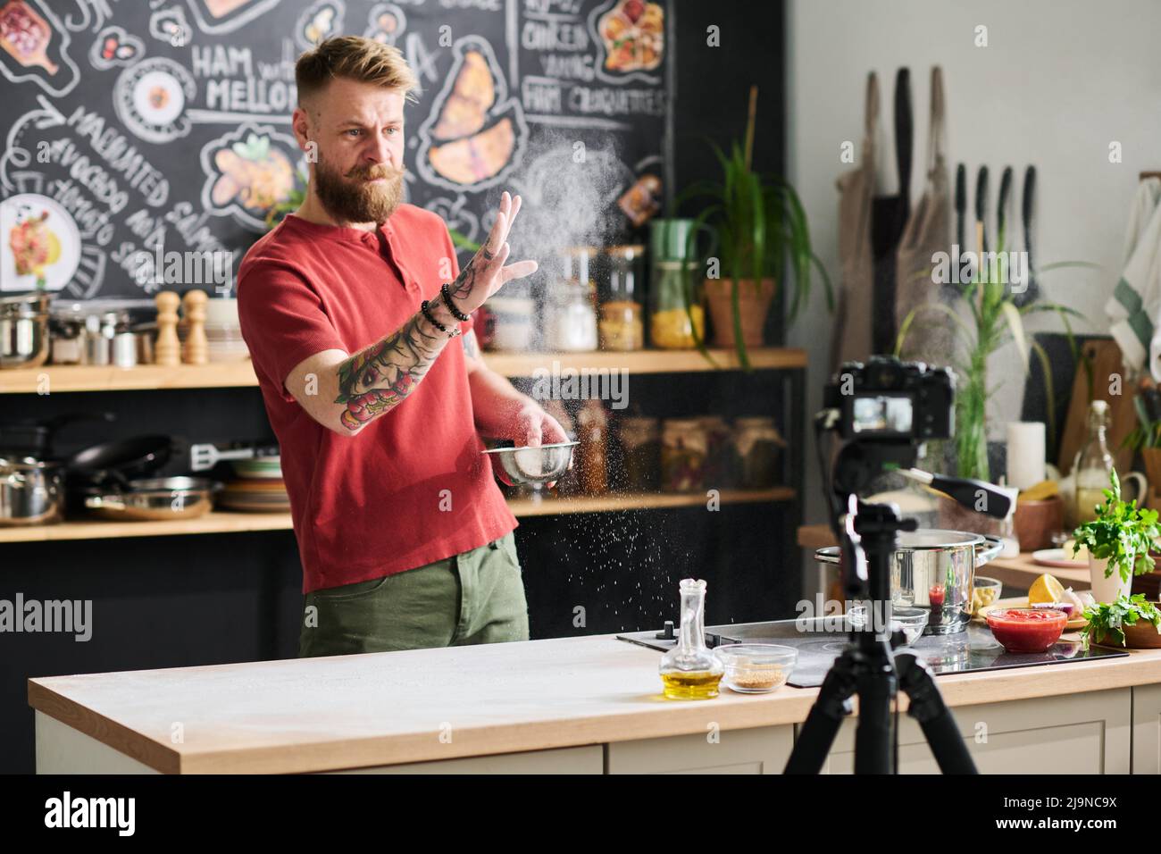 Stylish Caucasian food blogger making content for vlog dusting flour in air while preparing table surface to make dough Stock Photo