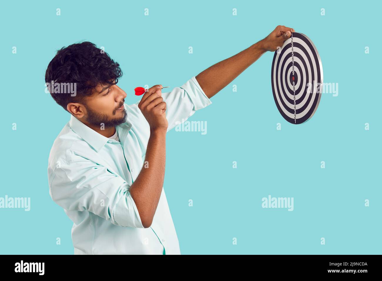 Side view of young Indian man holding target and aiming little arrow at bullseye Stock Photo