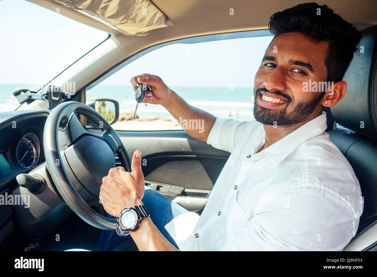 travel vacation happy indian man in white shirt collar buying new car and showing the key, sitting in car on beach sea india octan Goa .a trip to the Stock Photo
