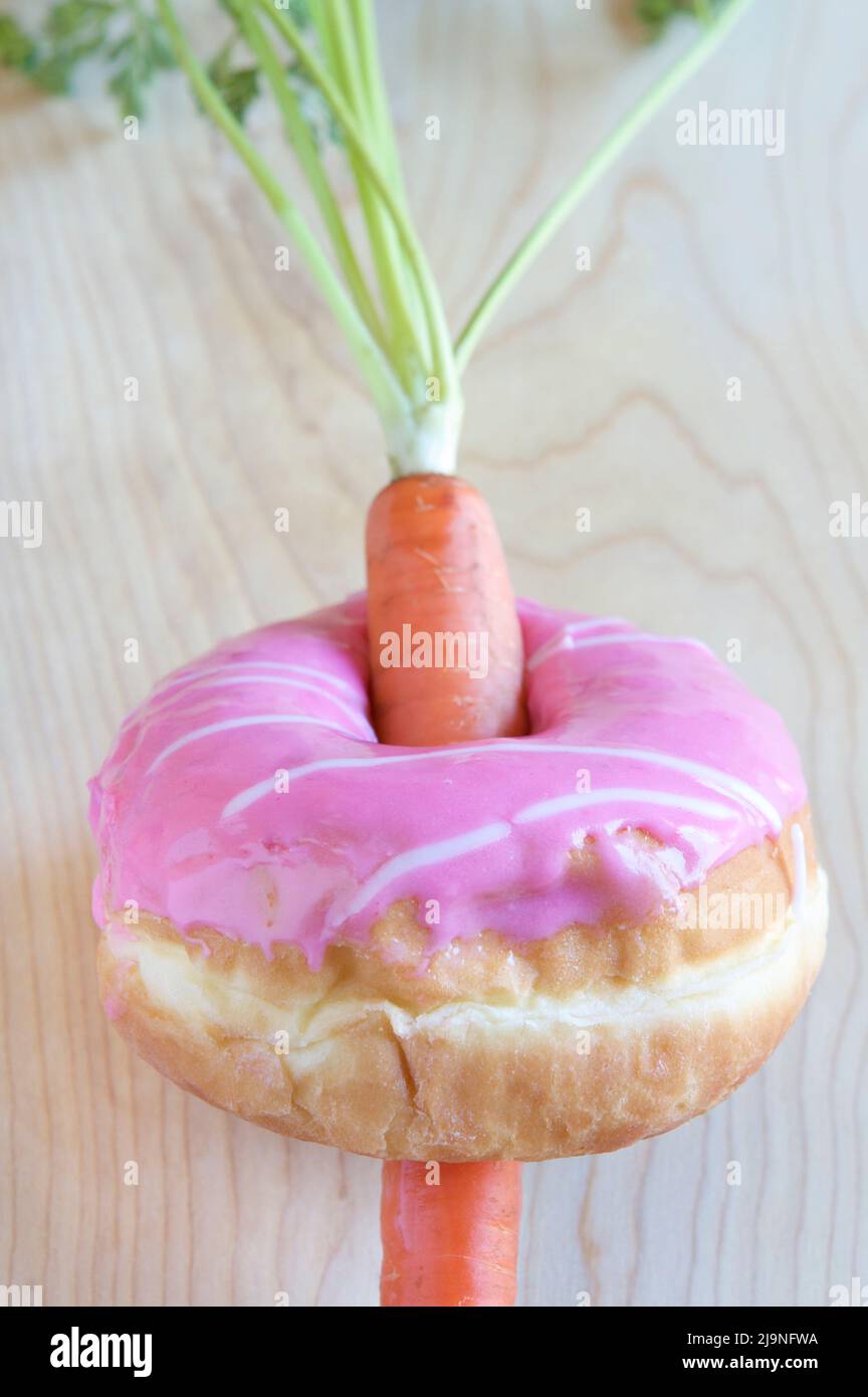 A single orange carrot with greens through the middle of a doughnut with pink frosting on wooden surface, creating a contrast between healthy and fat. Stock Photo