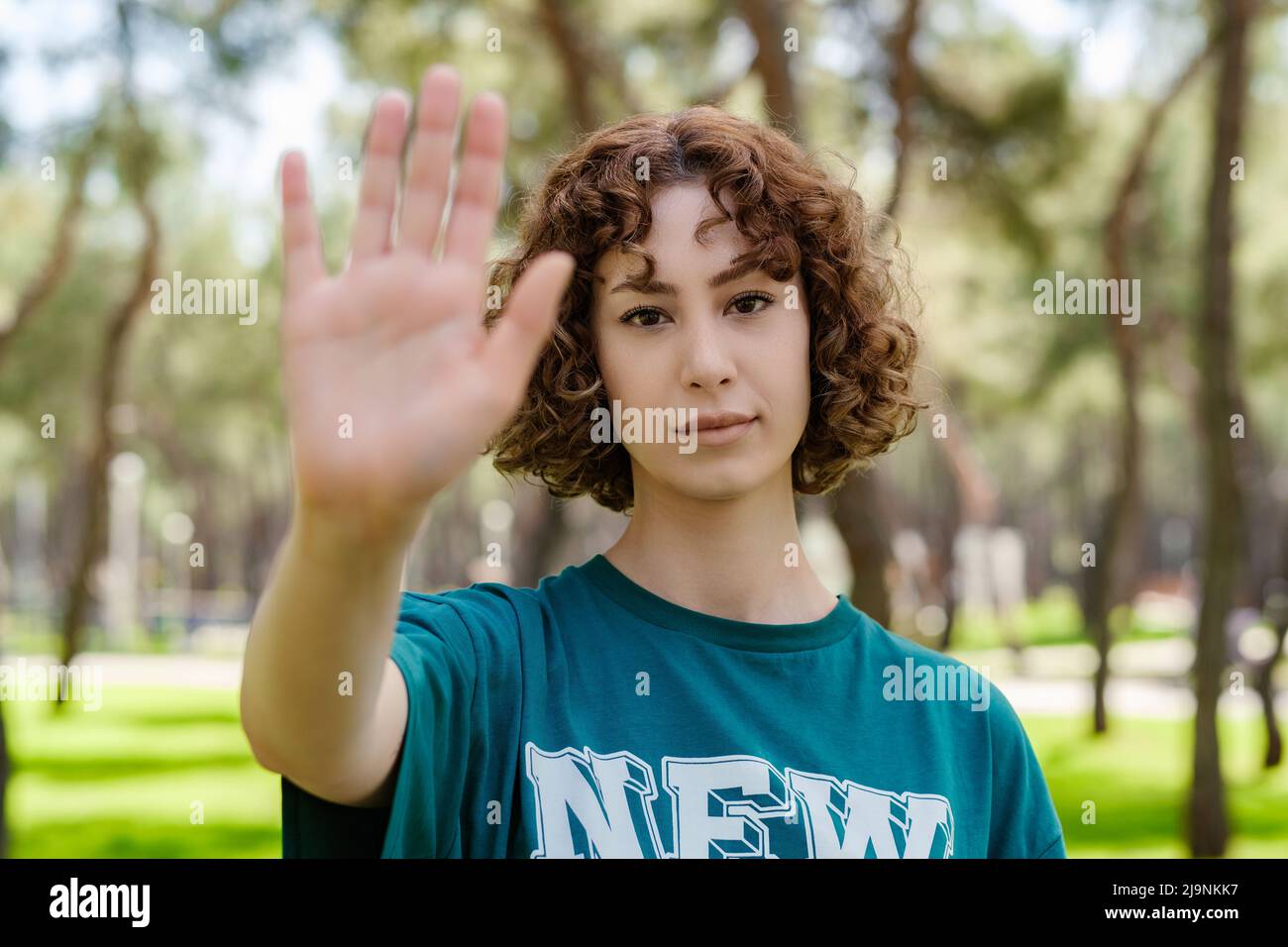 Young redhead serious woman wearing green t-shirt standing on city park, outdoors doing stop sing with palm of the hand. Warning expression with negat Stock Photo