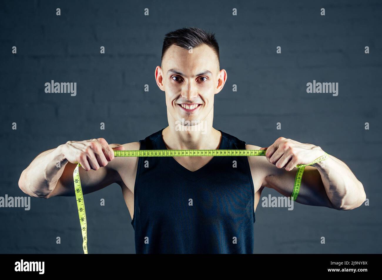 A young man measures his hand with a measuring tape in the gym on a dark background Stock Photo