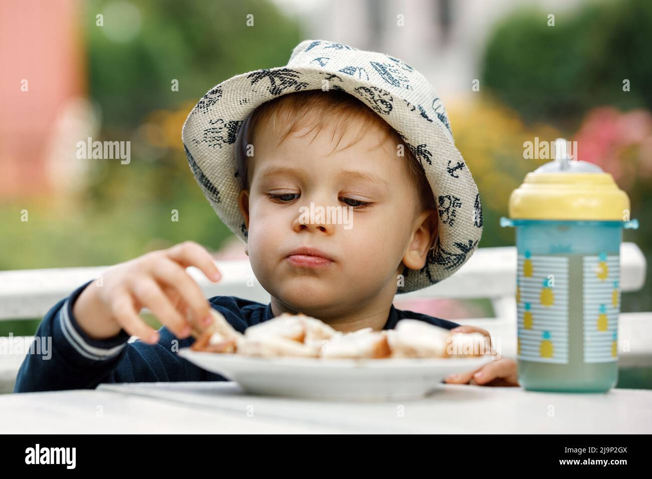 The boy eats toast for breakfast with juice in the background of a summer garden. A plastic cup with pineapple juice on the table. Stock Photo