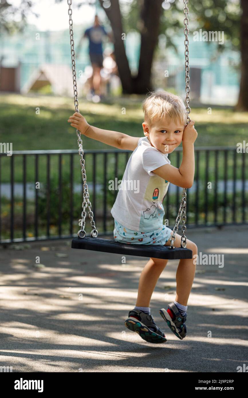 A mischievous little boy swings in a park on a chain swing, the child turns to the camera and smiles happily. Stock Photo