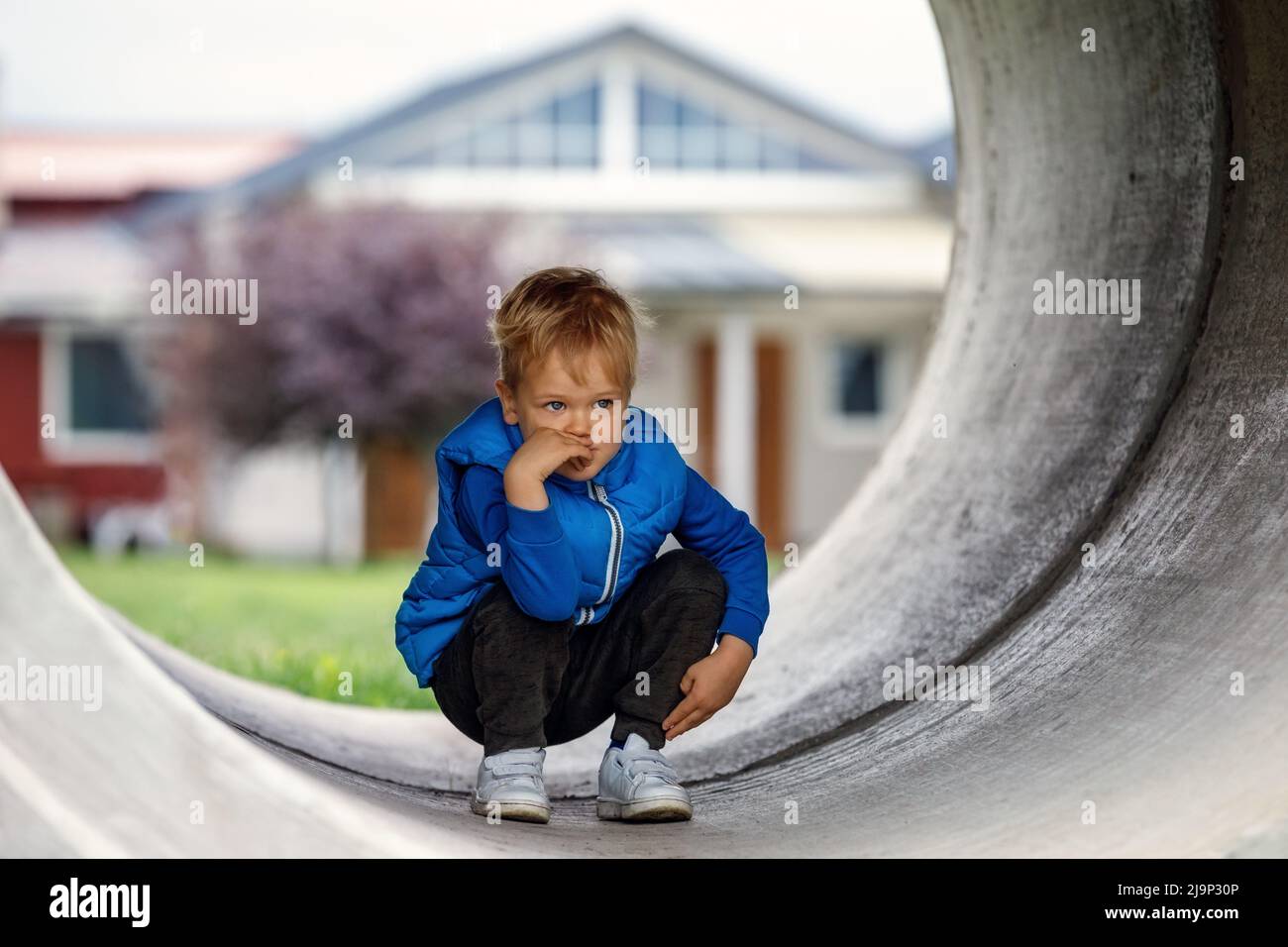 A cute kid wearing a blue vest squats in a concrete tunnel and thinks of something. Horizontal photo. Stock Photo