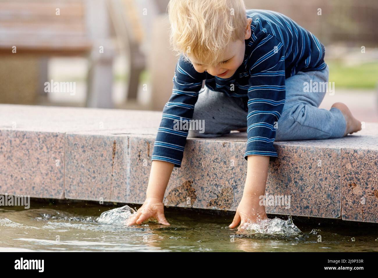Happy boy touching water in fountain. Boy rejoice in summer in a beautiful fountain. Active summer leisure for kids in the city. Stock Photo