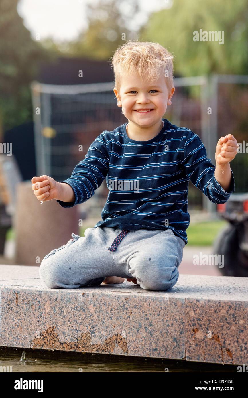 A little funny boy climbed on a parapet of a fountain and touch the water. Stock Photo