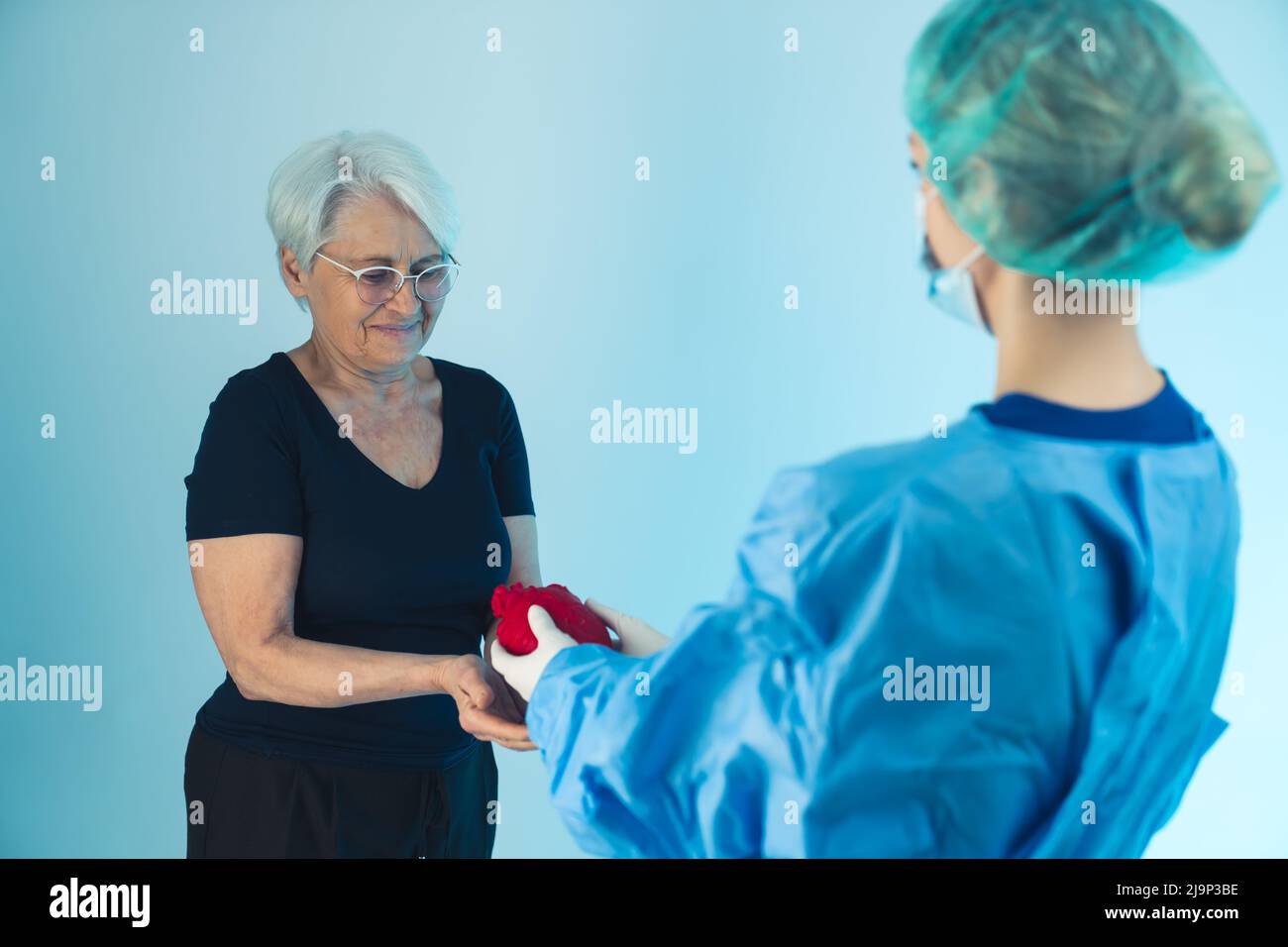 Female doctor in medical uniform, mask and medical cap giving an artificial human heart to an old lady with grey hair wearing a black outfit and glasses. Studio shot. High quality photo Stock Photo