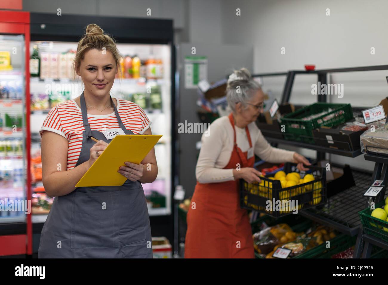 Young shop assistant in supermarket in vegetable shell, in bakcground is her colleague filling stock. Stock Photo